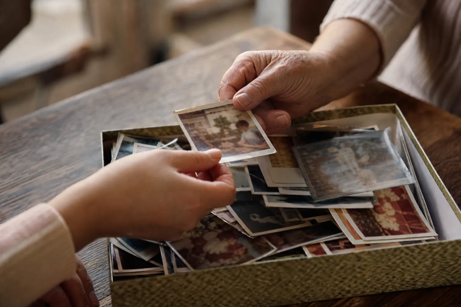 Two women's hands passing a faded snapshot over a shoebox of old family photos